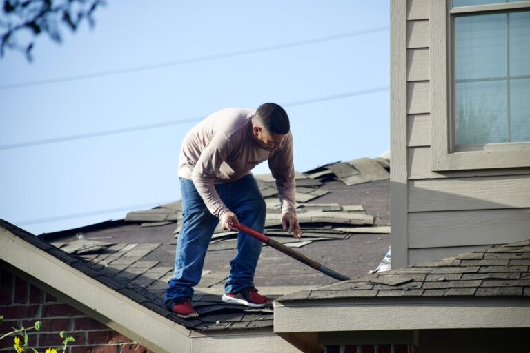 Workers removing old shingles with ladders and tarps set up around a house.
